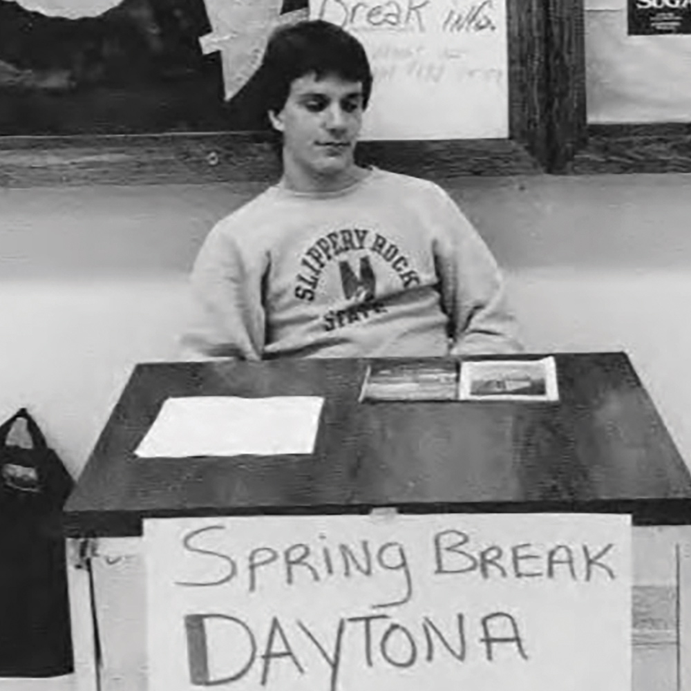 old photograph of a Slippery Rock student sitting at a table with a sign that reads “Spring Break Daytona”