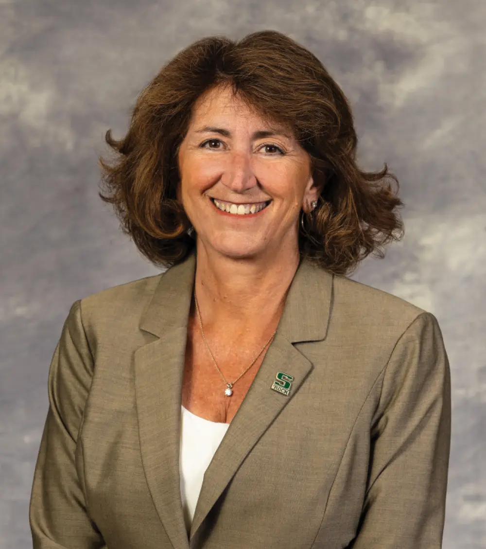 Portrait orientation close-up headshot photograph of Karen Riley smiling in a dark faded dull beige business blazer suit, white blouse top underneath, a thin chrome necklace, and an attached pin containing the green uppercase letter S plus beneath that the words ROCK which is attached to her left side of the business blazer suit that symbolizes her affiliated representation for Slippery Rock University