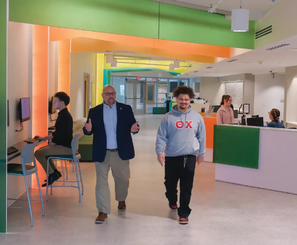 Portrait orientation indoor photograph of an older bald man presumably a campus professional individual in thin black prescription eyeglasses, dark navy blue business blazer suit, tan dress pants, and dark brown work dress shoes is visibly seen walking forward, gesturing with hands in air in motion as he is next to a smiling young man presumably a college student in a gray hoodie, black pants, and black/red basketball casual sneakers as they are located inside a modern, brightly lit indoor space with colorful walls and other people working or sitting in the background at the Campus Success Center at Slippery Rock University