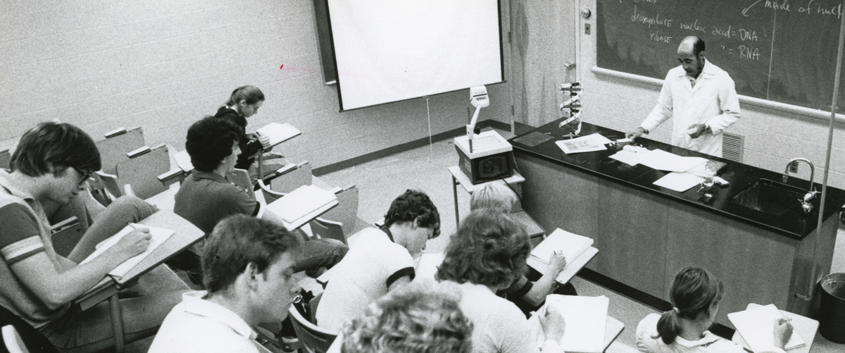 black and white high angle photo of Thomas Gaither in the middle of a lecture at the front of the lecture room as seated students diligently write notes