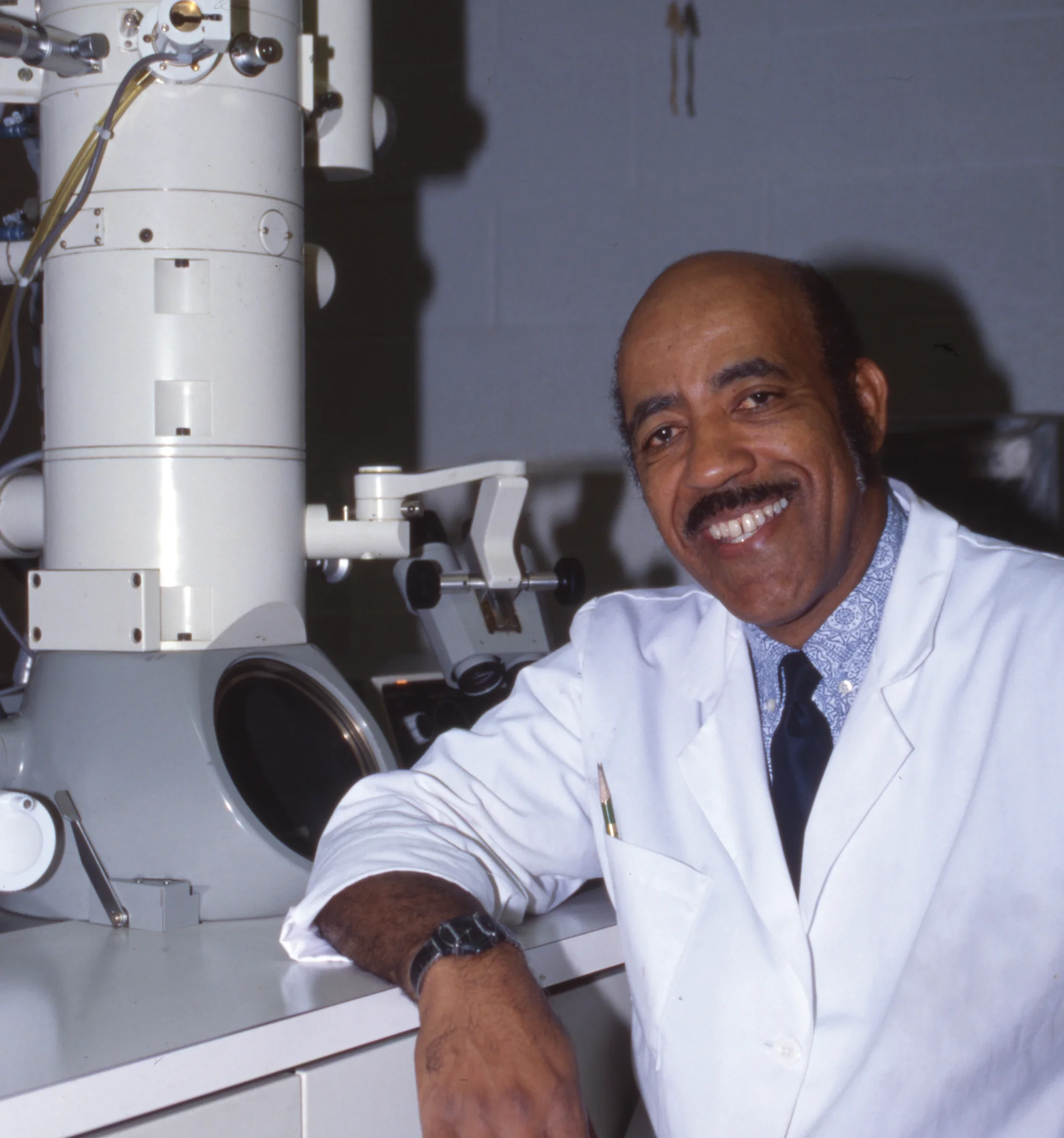 portrait of Thomas Gaither, wearing a white lab coat and leaning his elbow on a large machine