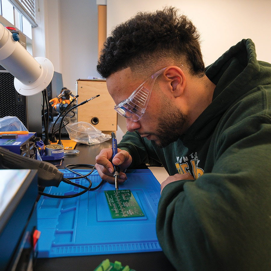 close cropped profile view of student Nicco Mickens wearing safety goggles and sitting at a lab desk working on the circuit board of an air heater he built