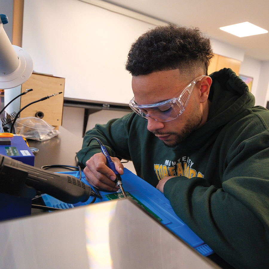 close cropped quarter view of student Nicco Mickens wearing safety goggles and sitting at a lab desk working on the circuit board of an air heater he built