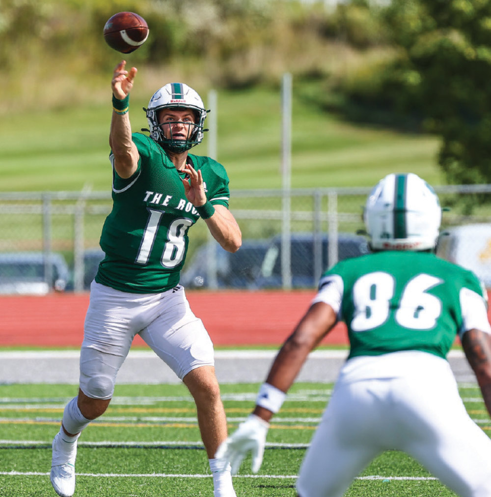 A football player, wearing a green jersey with the number '18', white pants, and a white and green helmet, is captured mid-throw on a football field