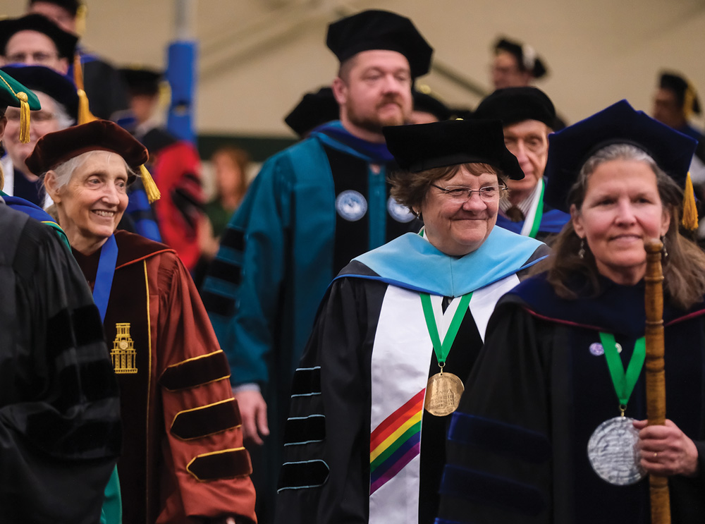 Academics in ceremonial robes at a formal event, with medallions and caps, one holding a staff.