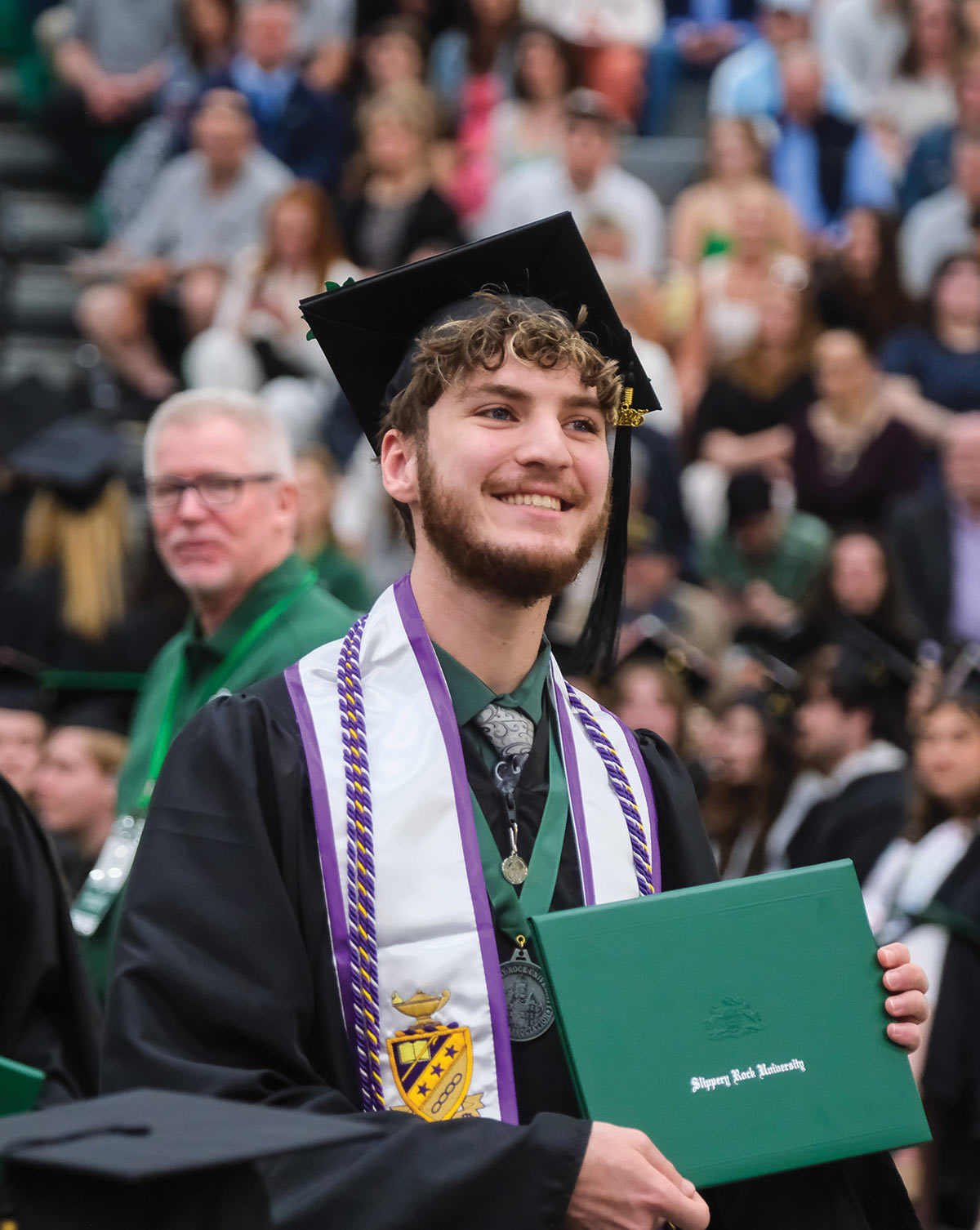 A graduate at a ceremony holding a diploma cover, smiling, with a crowd in the background.