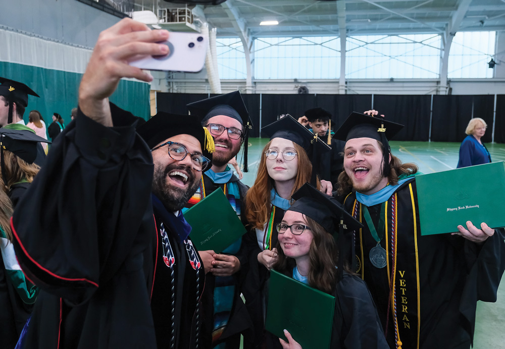 Graduates taking a selfie at a commencement ceremony indoors, wearing black gowns and caps.