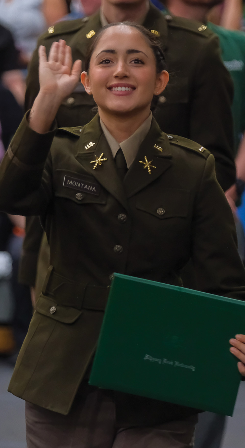 Person in a military-style uniform smiling and waving, holding a green folder.