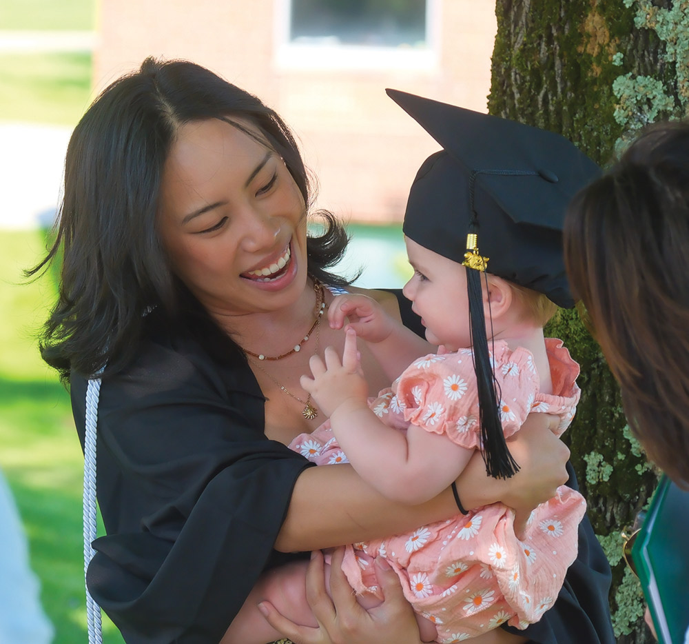 A woman in a graduation gown holds a baby wearing a graduation cap and floral dress, smiling warmly at each other.