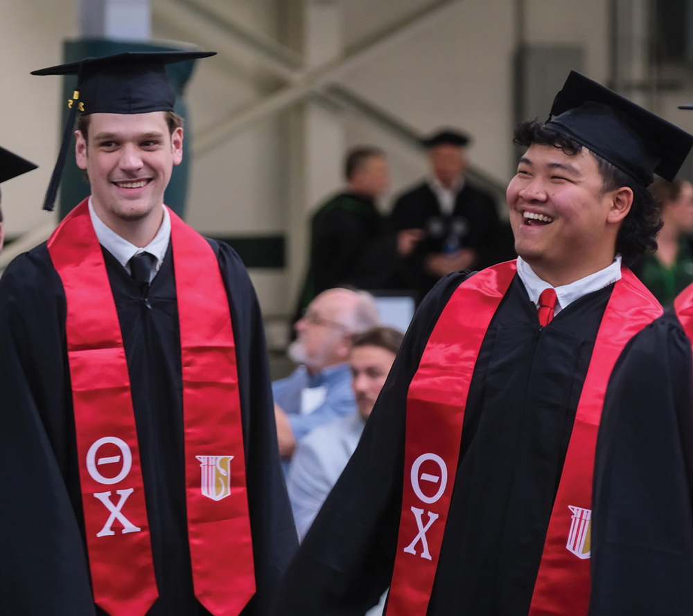 Two graduates in caps and gowns, smiling broadly with red stoles featuring Greek letters.