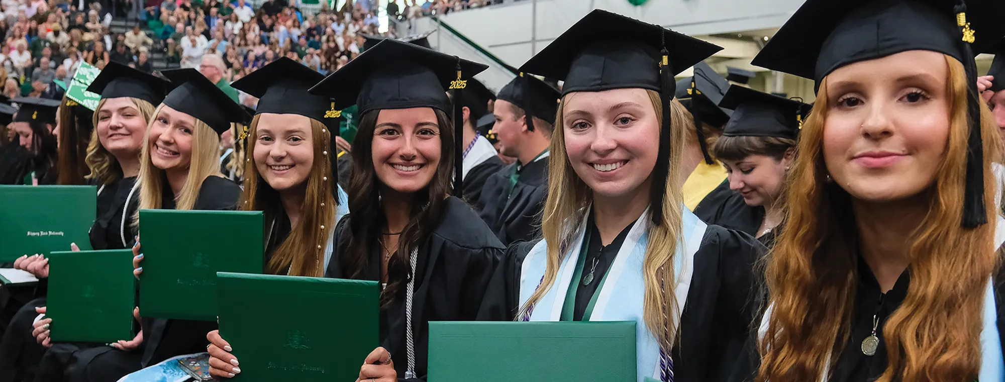 Graduates seated in a row, wearing caps and gowns, holding green diploma covers.