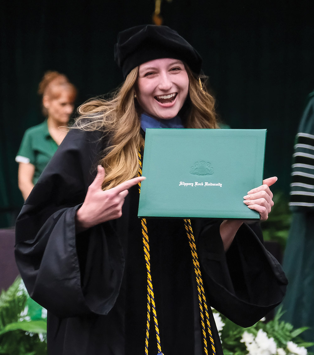A person in a graduation cap and gown smiles and holds a green diploma cover.
