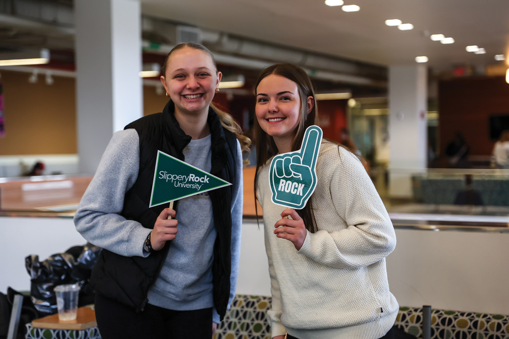 Two people smiling indoors, one holding a Slippery Rock University pennant, the other holding a foam finger.