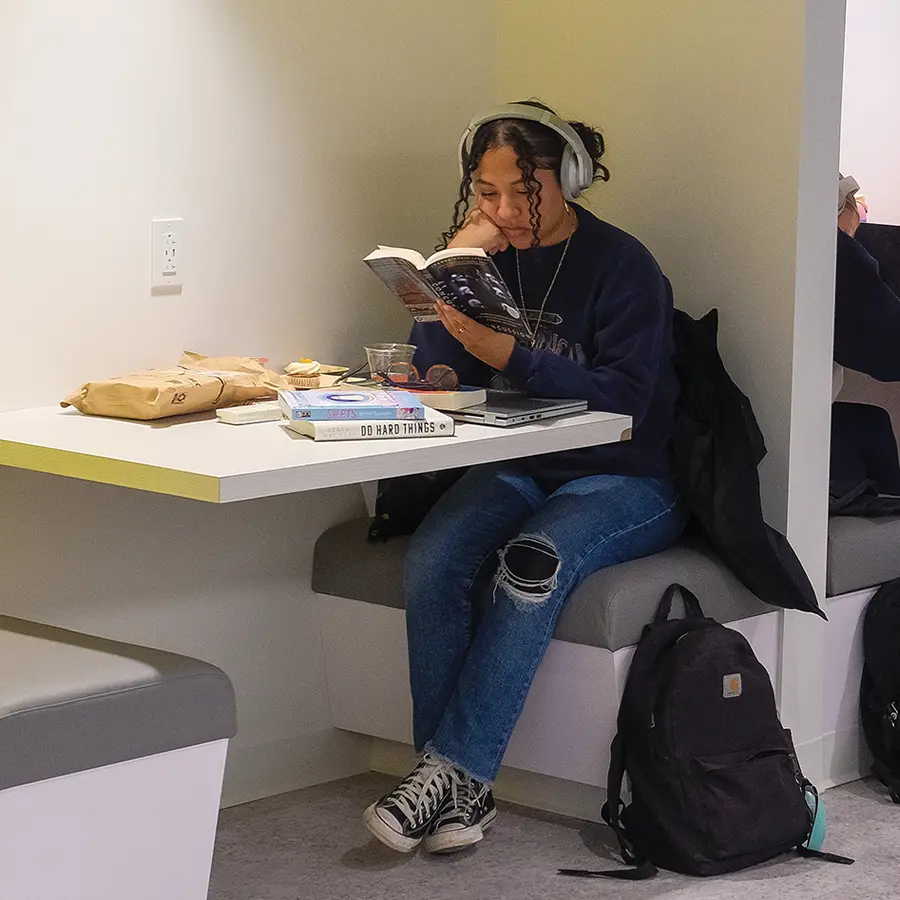 student sitting at a booth and wearing headphones while reading a book