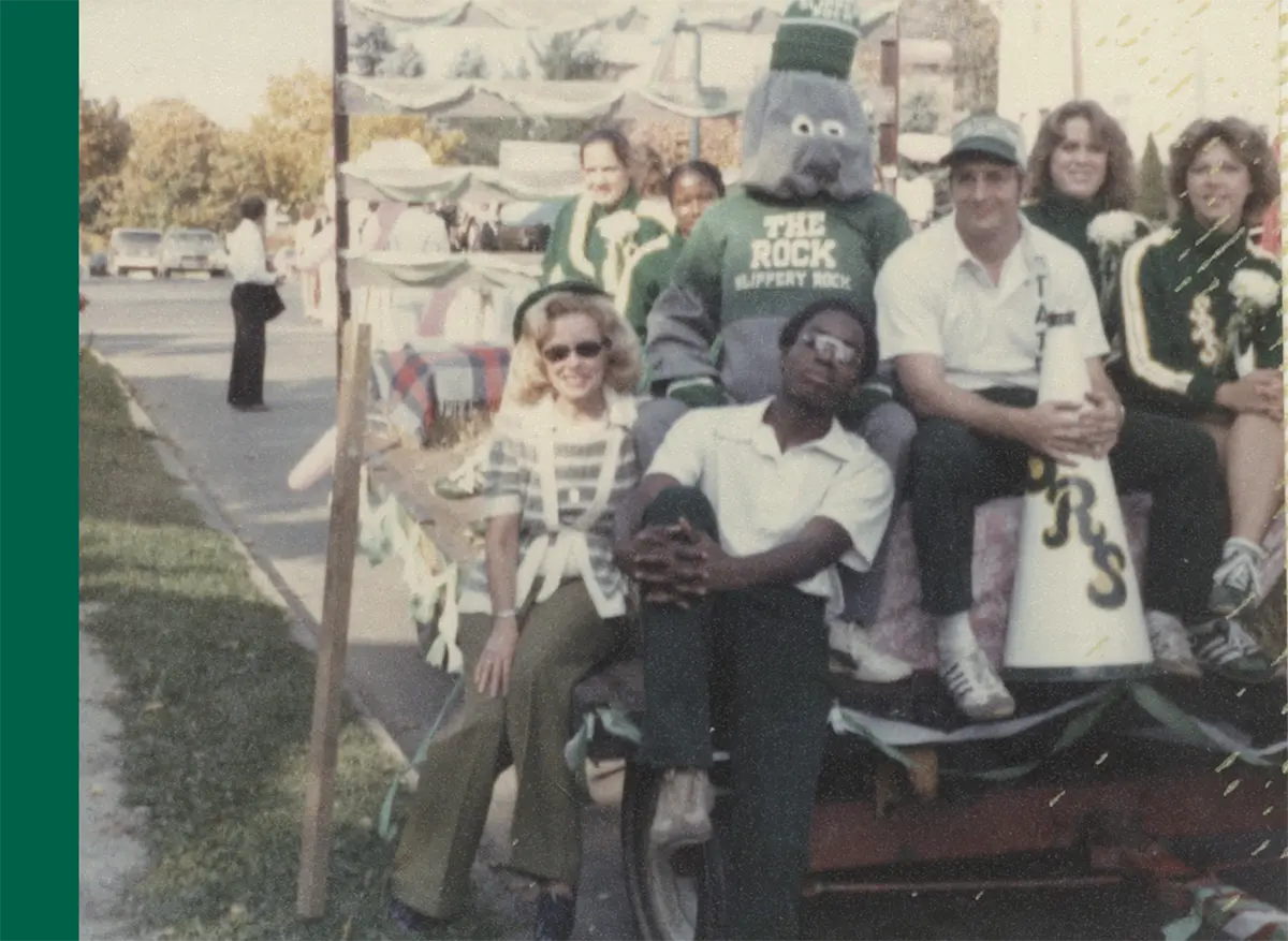 A vintage photo of a group of people and the former Slippery Rock University (SRU) mascot, a gray rock-like figure, riding on a parade float or vehicle of some kind; The mascot is wearing a dark green graphic t-shirt that says THE ROCK SLIPPERY ROCK and a beanie that says SLIPPERY ROCK and a large white megaphone labeled S R S is visible which is seen held by a random man who is a part of the group