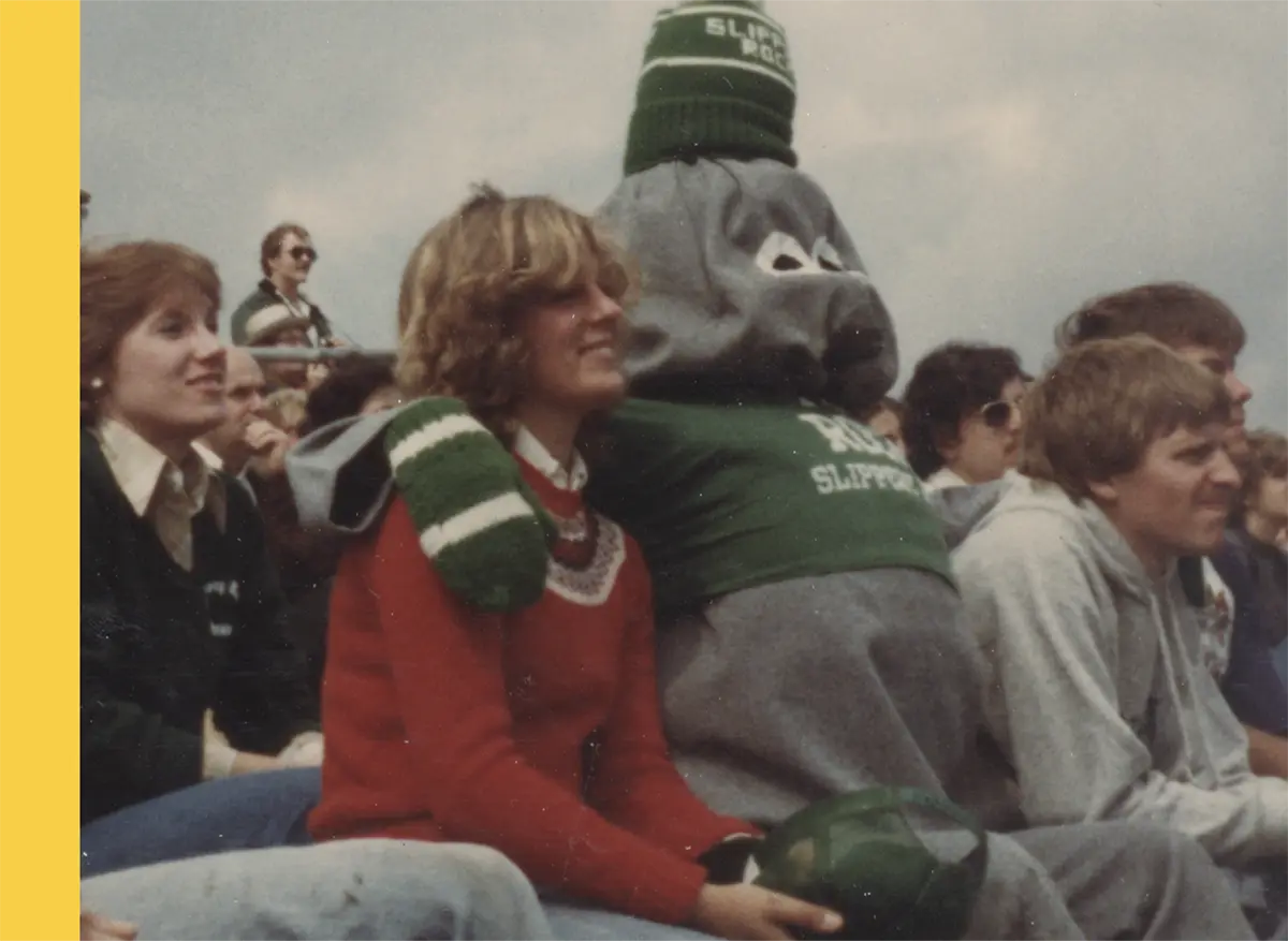 A vintage photo of spectators in a stadium; In the foreground, a woman in a red sweater holds a green object presumably a hat and a man in a gray hoodie sits next to her; Behind them, the former Slippery Rock University (SRU) mascot is wearing a gray rock-like mascot costume with a green graphic t-shirt that says ROCK SLIPPERY ROCK and a dark green striped beanie that says SLIPPERY ROCK