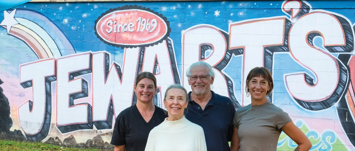 A group photo of four people standing in front of a colorful wall mural for "J.E. WART'S Since 1969."