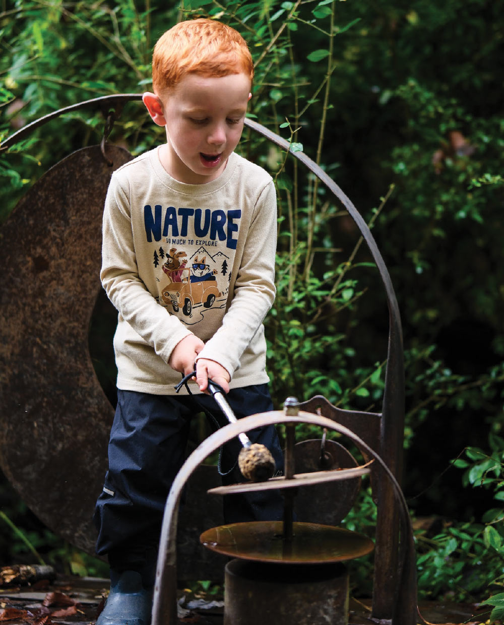 A young boy plays a rusted outdoor metal musical instrument in a garden.