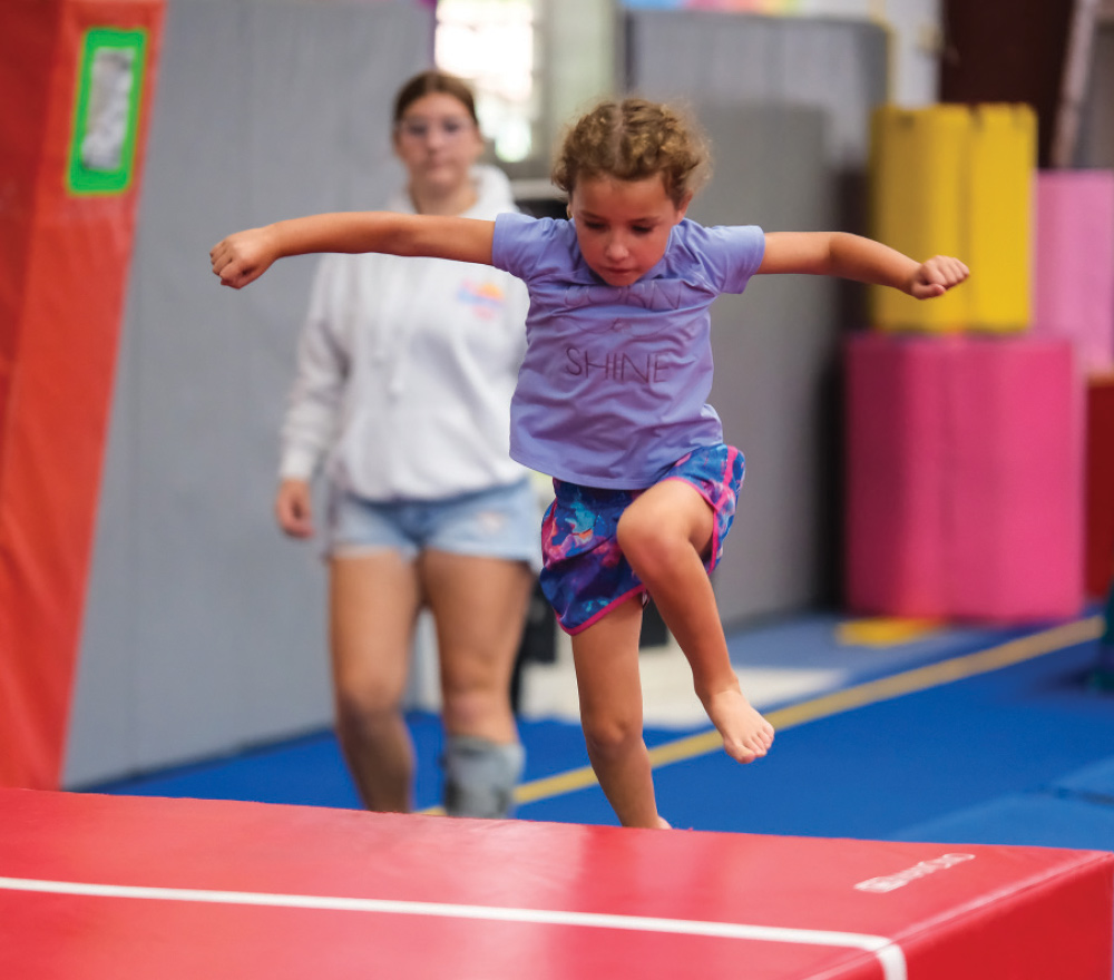 A young girl in a purple shirt and patterned shorts jumps barefoot off a red mat in a gym.