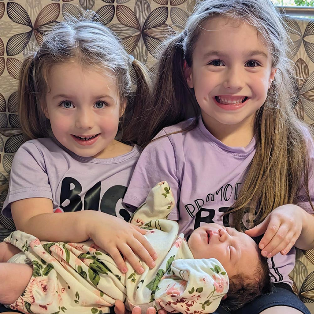 Two young girls with pigtails wearing matching purple shirts smile while holding a sleeping newborn baby dressed in a floral outfit on a patterned chair.