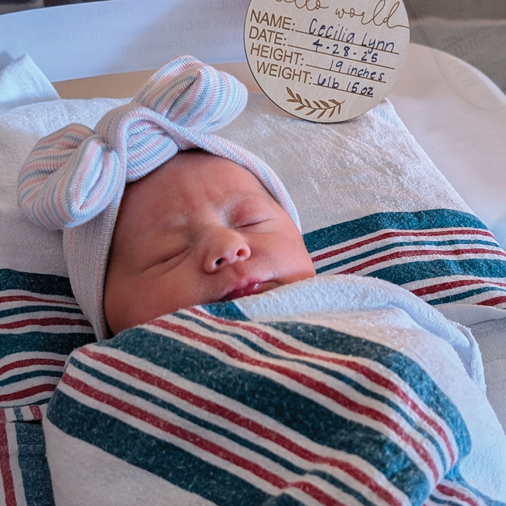 A sleeping newborn baby swaddled in a striped hospital blanket wearing a large knit bow, lying next to a wooden announcement sign.