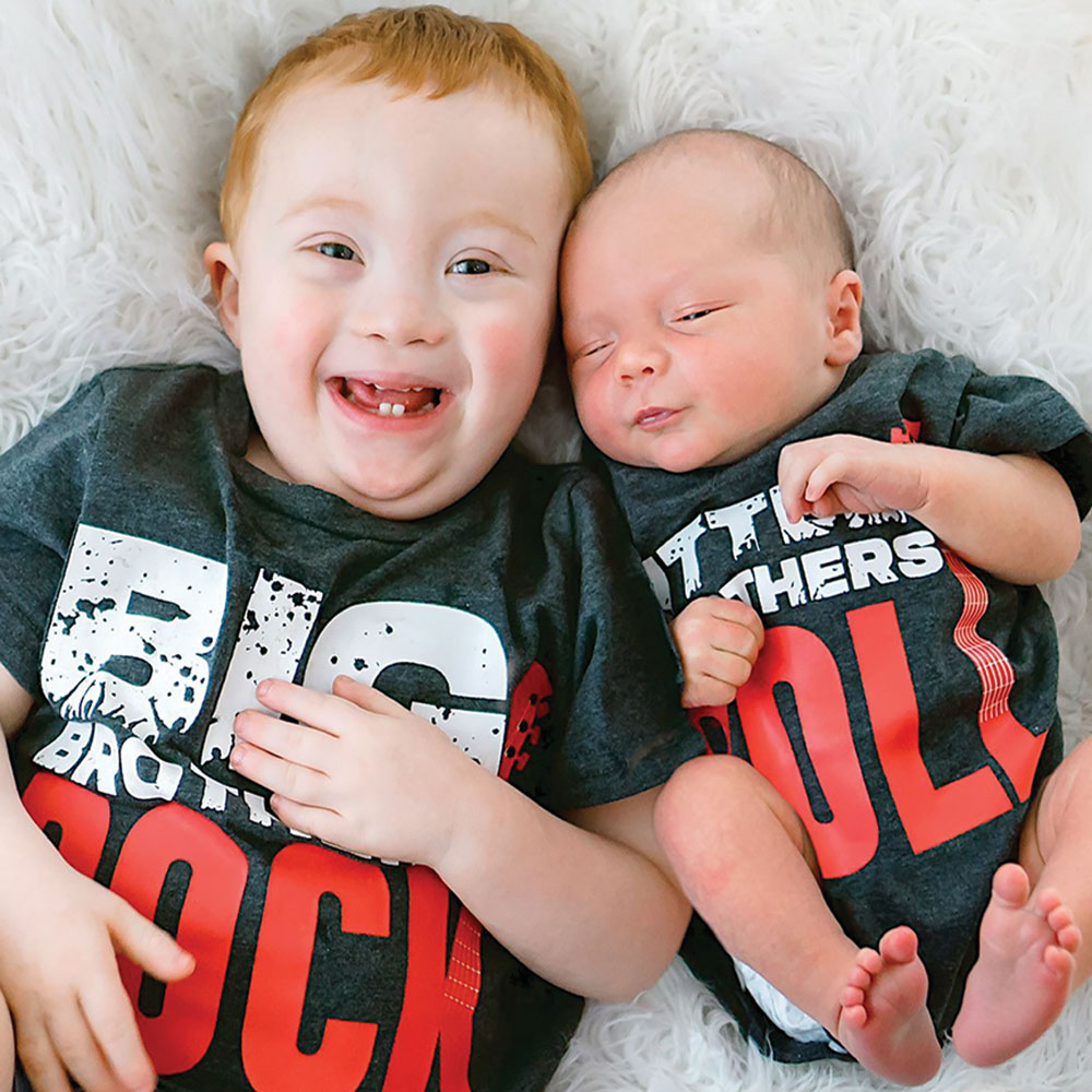 A young boy with red hair smiles broadly while lying on a fluffy white rug next to a newborn baby, both wearing matching dark grey 
