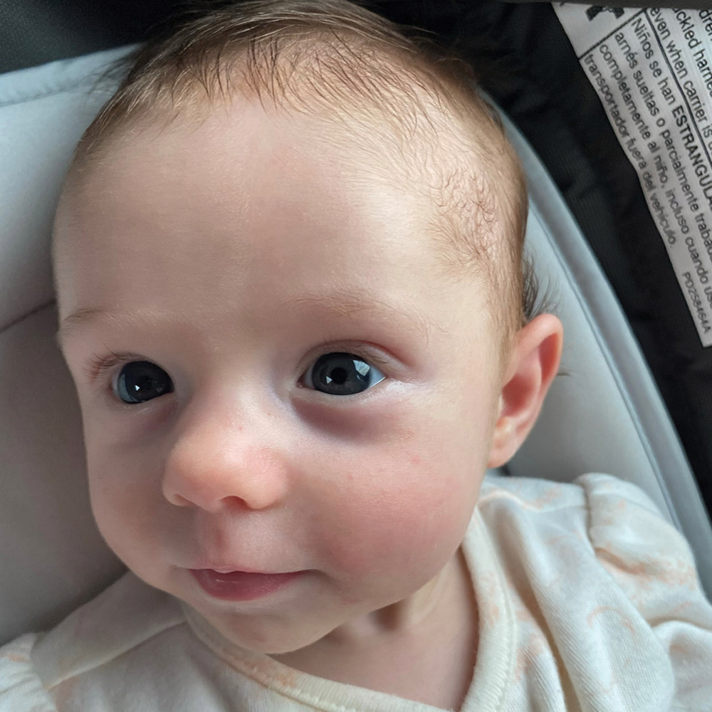 A close-up of an alert baby with light wispy hair and wide blue eyes looking slightly to the side, resting in a grey padded car seat.