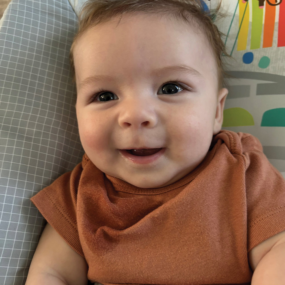 A close-up portrait of a happy baby with light brown hair and dark eyes wearing a rust-colored t-shirt, smiling while sitting in a patterned baby seat.