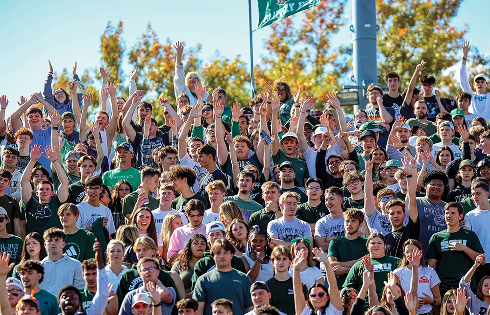 A packed student section fills the stadium stands on a sunny day. The enthusiastic crowd wears green and white school colors, with many students raising their hands to cheer for the team.