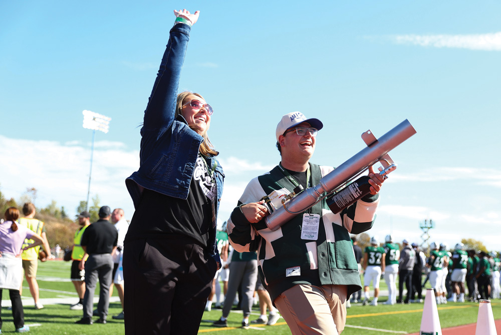 A smiling woman in sunglasses raises her arm in excitement next to a man holding a large silver t-shirt cannon. They walk along the sidelines of the football field, engaging with the crowd.