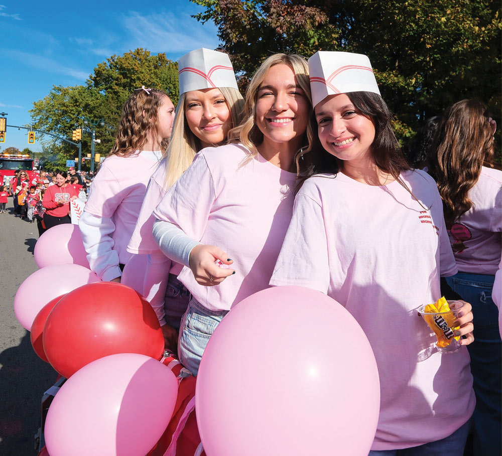 Three smiling students participate in the homecoming parade, wearing matching pink t-shirts and retro paper diner hats. They stand behind pink and red balloons on a float on a sunny day.