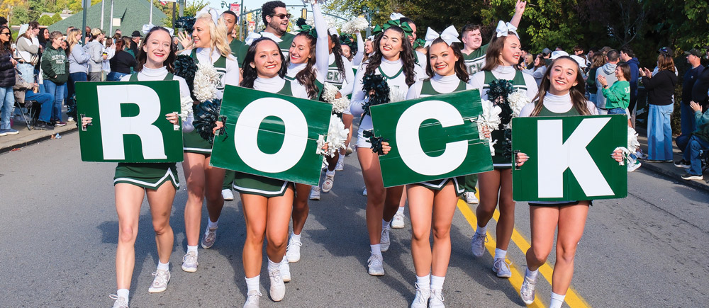 Cheerleaders in green and white uniforms march down a street during a parade. The front row holds large green placards with white letters, collectively spelling out "ROCK" to the crowd.