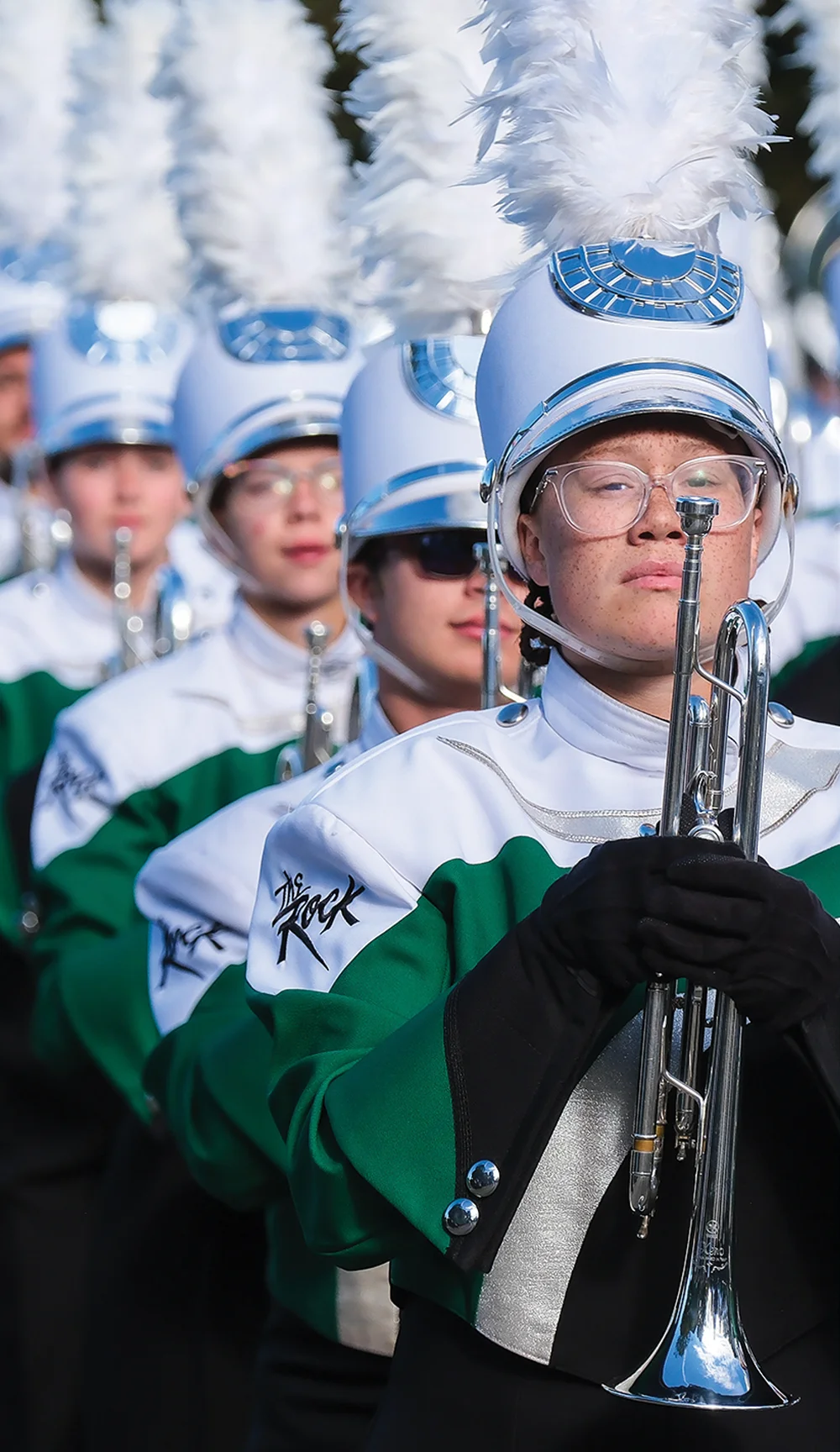 A uniformed marching band member holding a silver trumpet in the foreground, wearing glasses and a white plumed shako. Other band members in green and white uniforms stand in formation behind.
