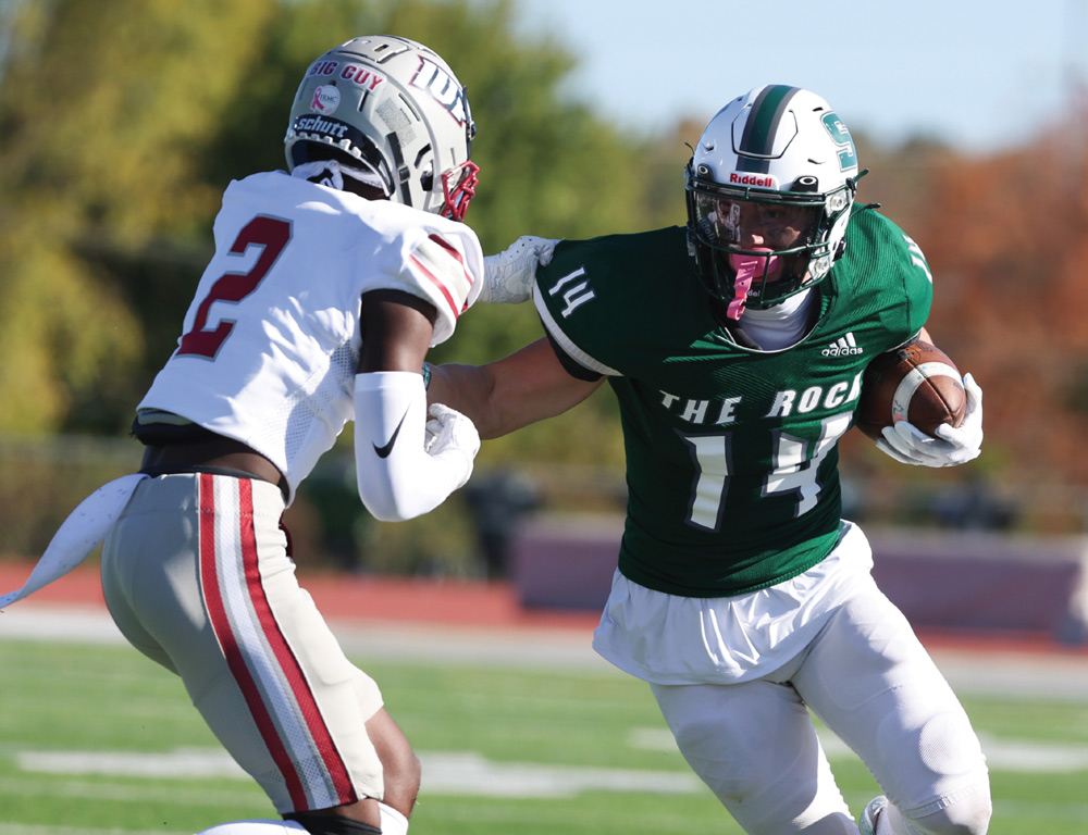 A football player in a green "The Rock" jersey runs with the ball, bracing for a tackle from an opposing player in white. The action takes place on a bright, sunny field during the game.