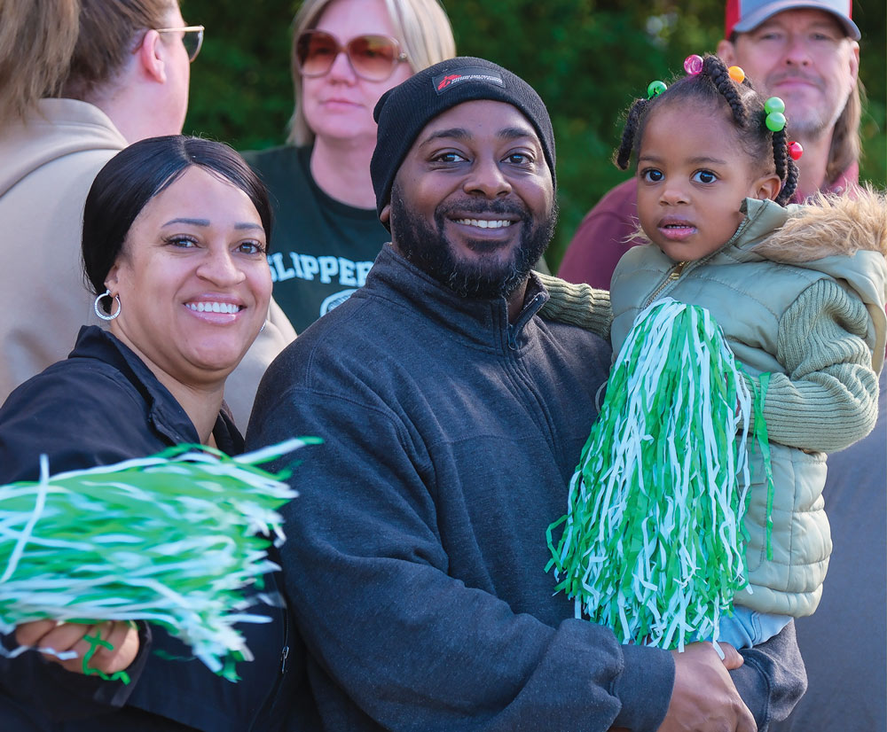 A smiling family attends the homecoming festivities. A man holds a toddler wearing a green puffy vest, while a woman beside him holds a green and white pom-pom, enjoying the event.