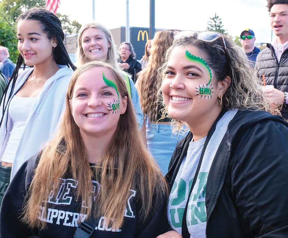 Two smiling female students pose for a photo with matching green face paint designs featuring swirls and "SRU" lettering on their cheeks. They stand in a crowd of homecoming attendees.