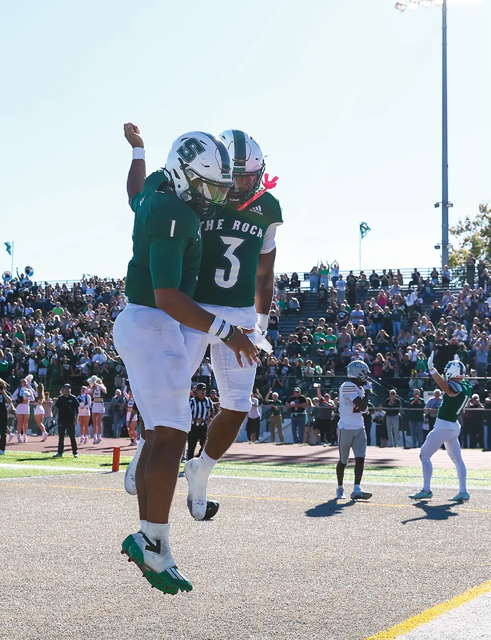 Two Slippery Rock football players, wearing green jerseys numbered 1 and 3, jump in the air to celebrate a play. Fans cheer in the background stands under a clear blue sky.