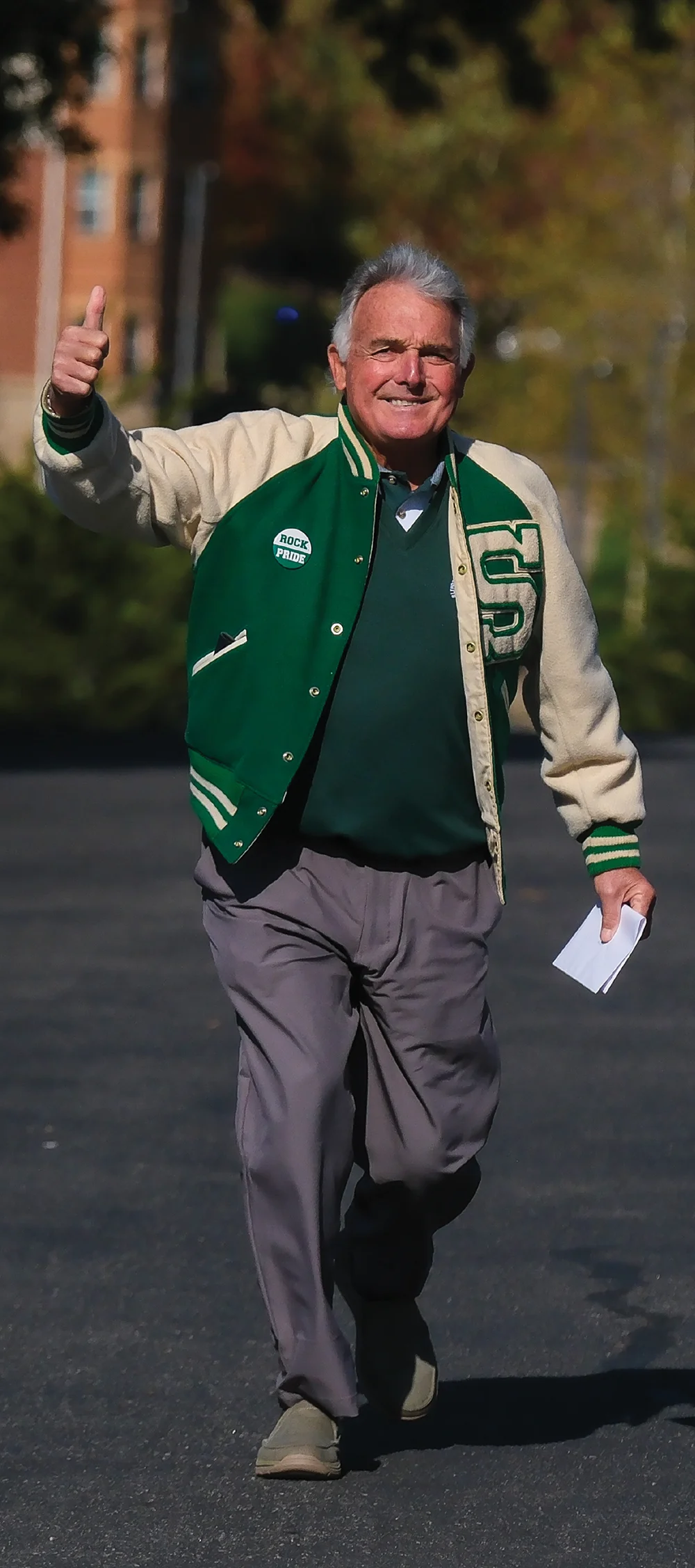 An older man walks toward the camera with a big smile, giving a thumbs-up gesture. He wears a green and white varsity letterman jacket with an "S" patch and a "Rock Pride" button.