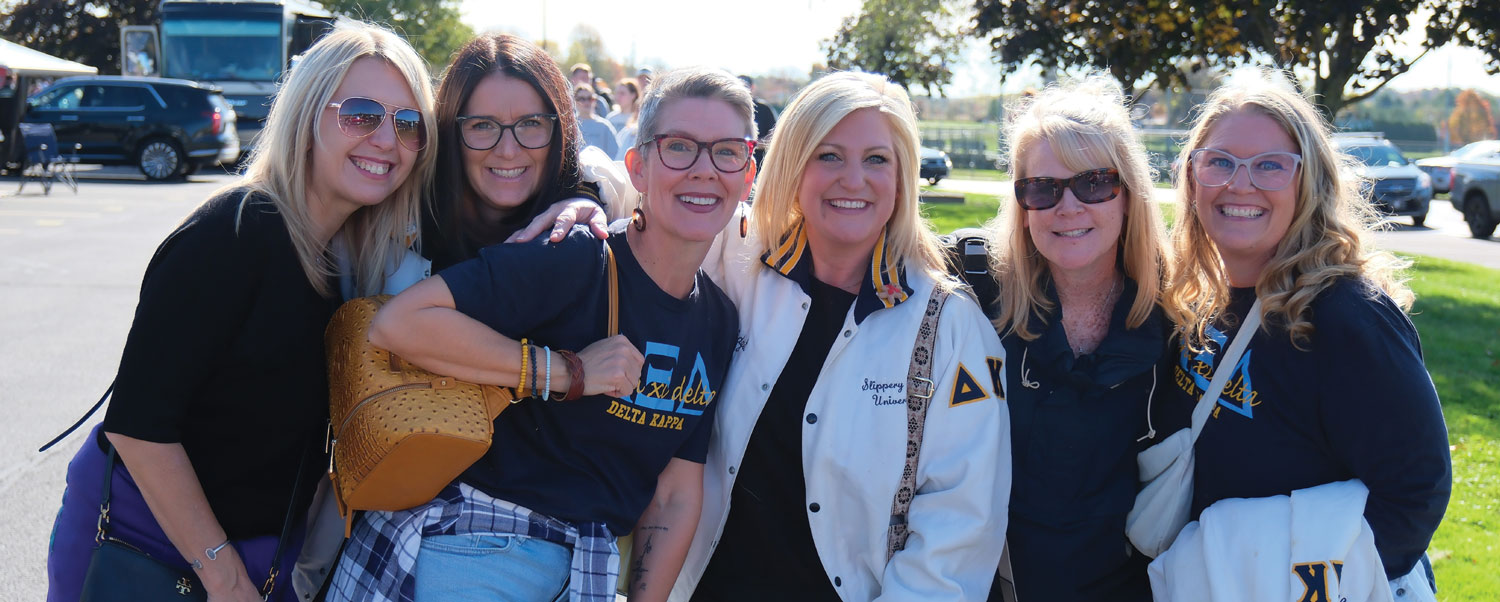 A group of six smiling women pose together outdoors in a parking lot. Several wear matching navy blue t-shirts with yellow Greek letters, and one wears a white Slippery Rock jacket.