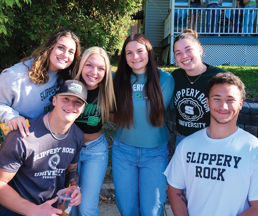 A group of six smiling students poses together outdoors on a sunny day. They wear assorted Slippery Rock University t-shirts and sweatshirts, standing on a sidewalk near a house porch.