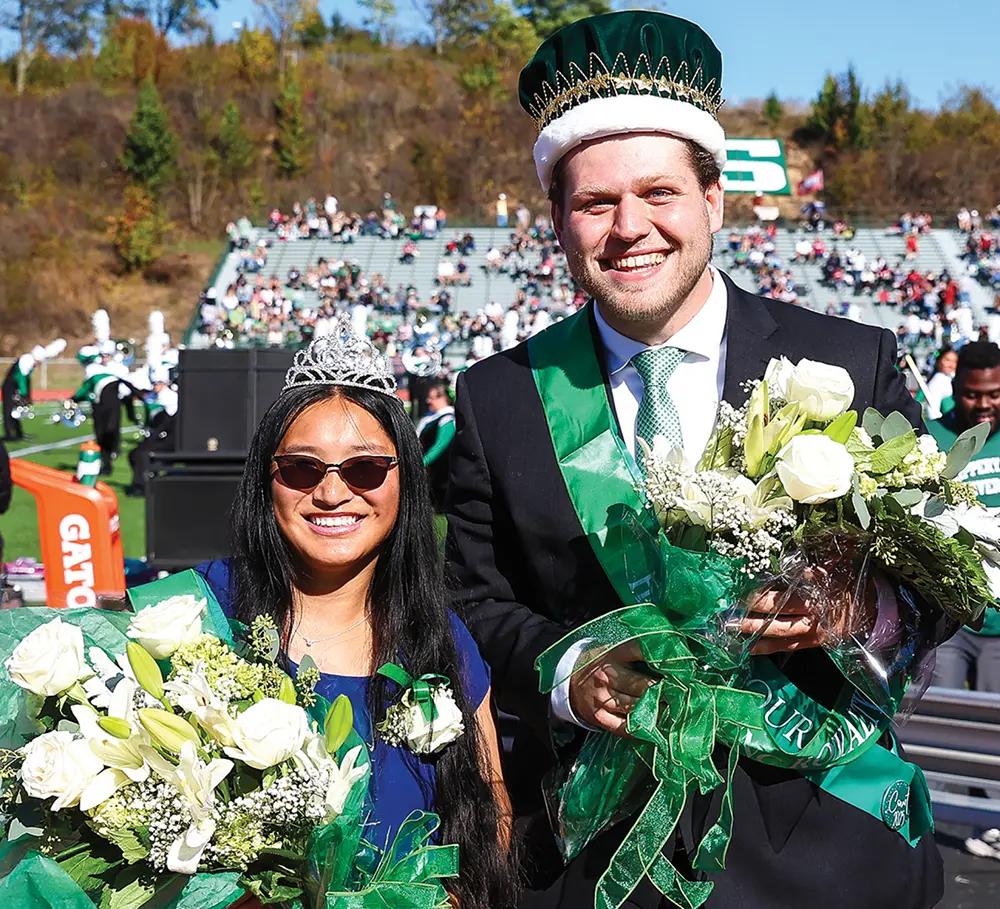 A smiling Homecoming King and Queen pose on the football field. The King wears a green sash and crown, while the Queen wears a tiara and sunglasses; both hold large bouquets of white roses.