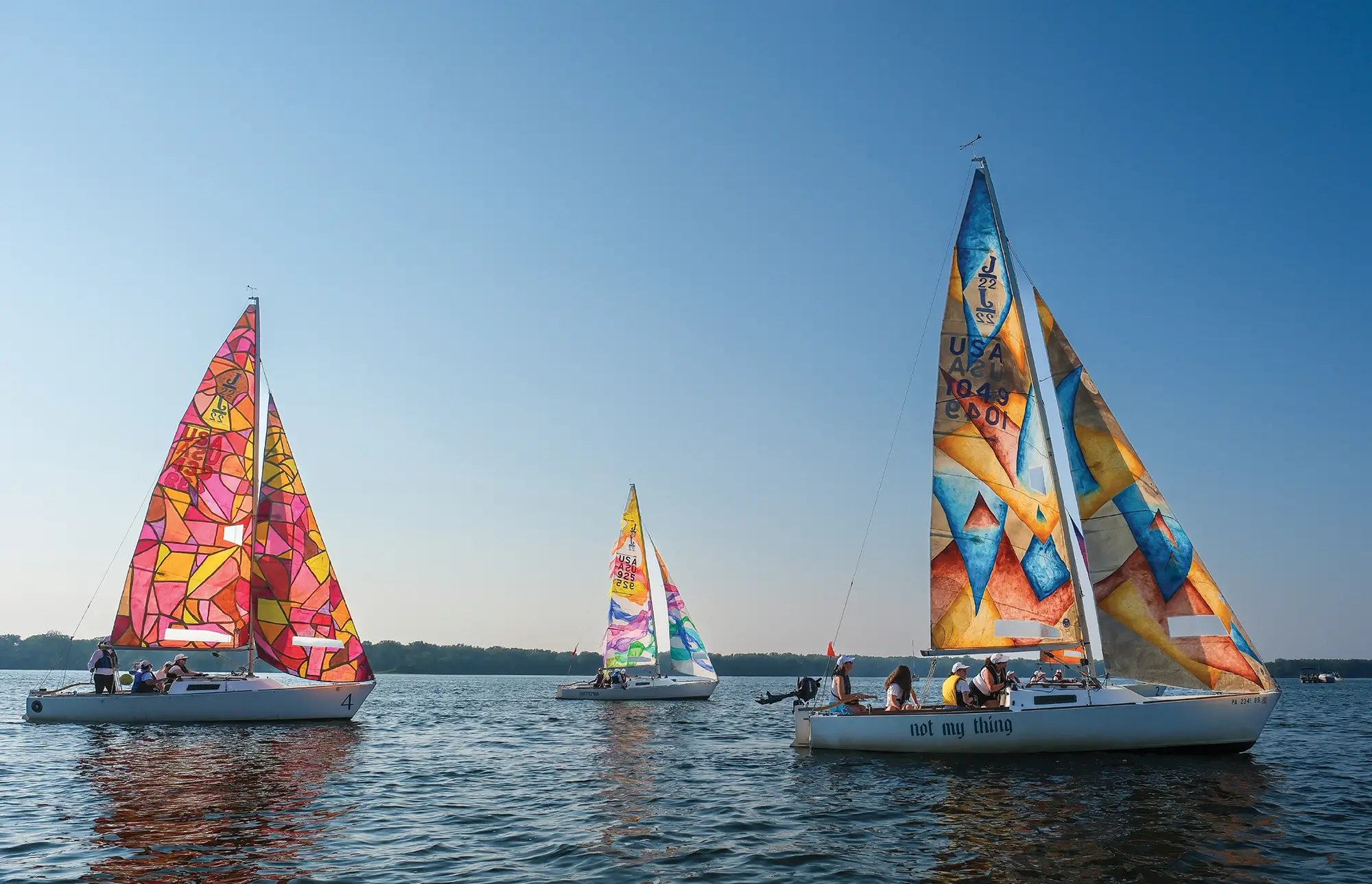Sailboats with colorful geometric patterned sails on a calm body of water under a clear sky.