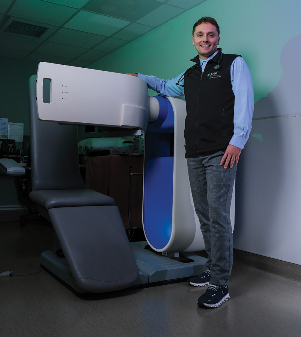 Man standing next to a gray and white medical device with a curved design and padded seat.