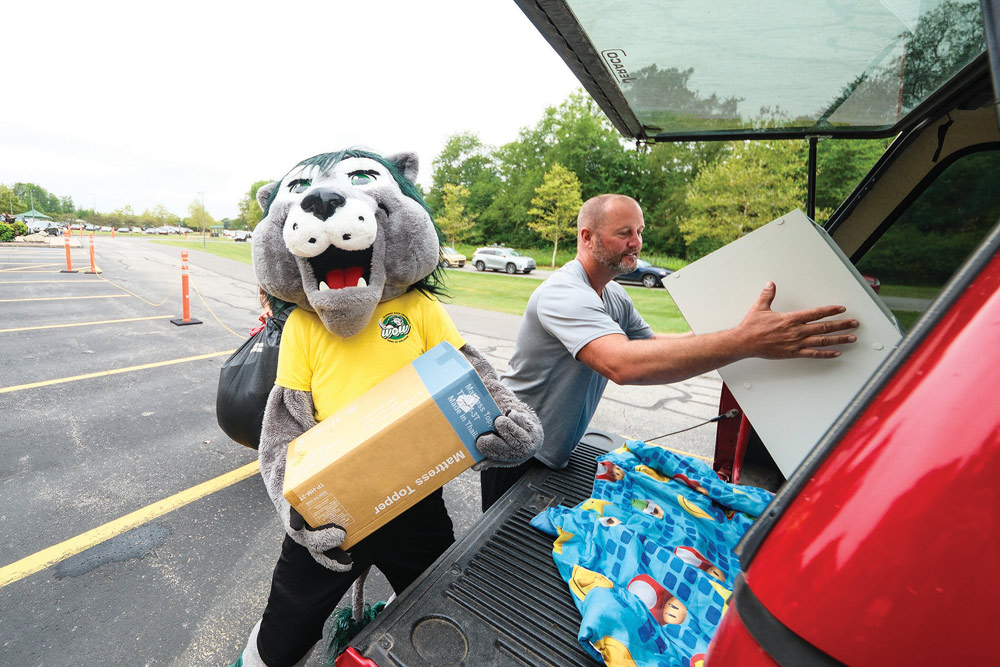 Slippery Rock University (SRU) mascot Rocky the Lion, a gray lion figure with dark green hair, is seen wearing a yellow graphic t-shirt and holding a box labeled Mattress Topper; Rocky is standing next to a man who is loading a white flat item into the back of a red pickup truck on SRU's move-in day