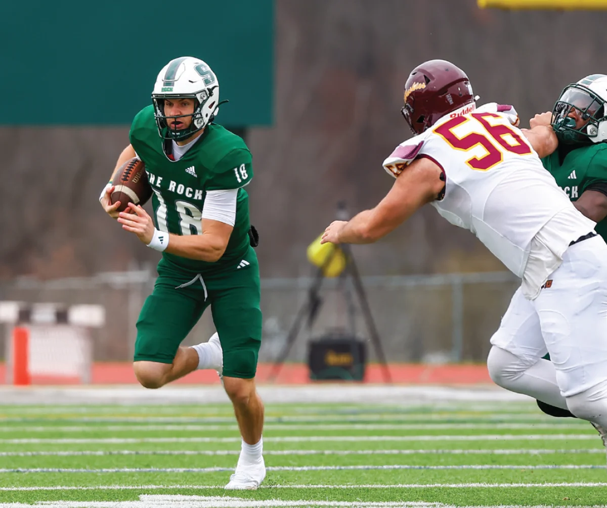 College football player in a green and white uniform (number 18) running down the field, evading a tackle from an opposing player