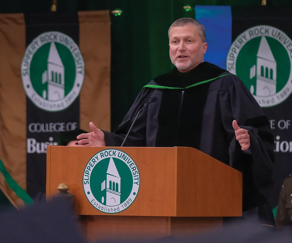 Keynote speaker addressing a large audience during a university event, with banners for the College of Business and Education visible.