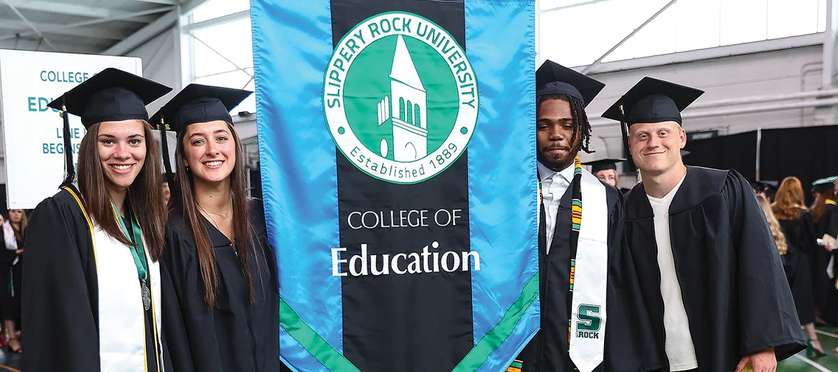 Four Slippery Rock University College of Education graduates in caps and gowns, celebrating commencement next to the school banner.