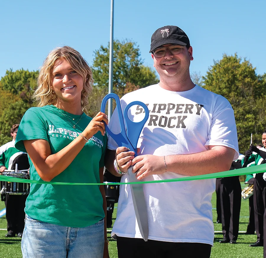 Man and woman in Slippery Rock 'The Rock' branded shirts cutting a green ribbon, marking the grand opening of a new campus addition.