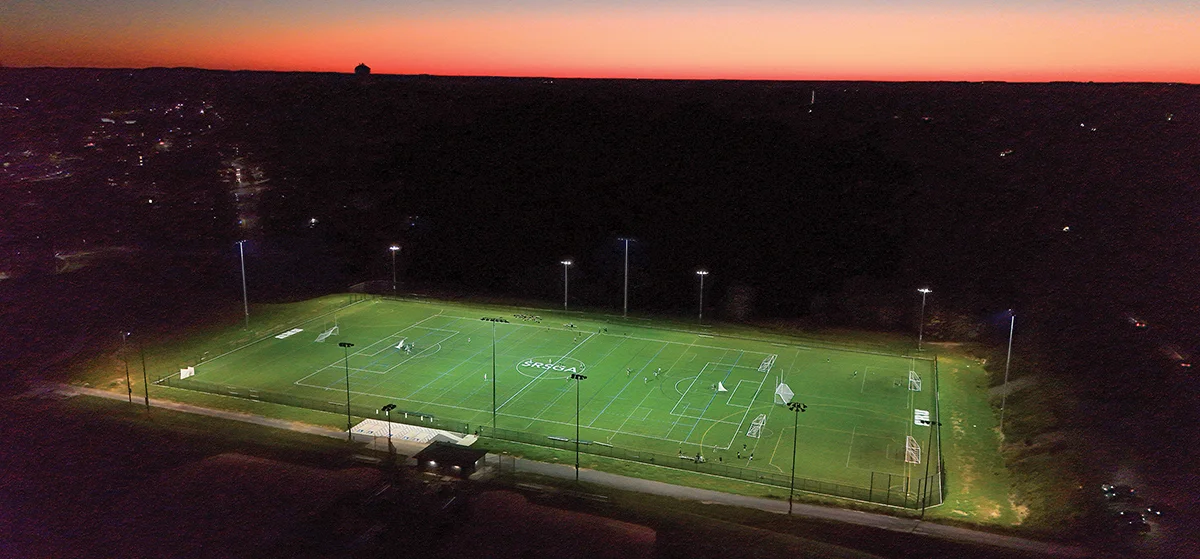 Aerial view of a brightly lit college soccer or turf field at night, showing the entire complex under tall stadium lights.