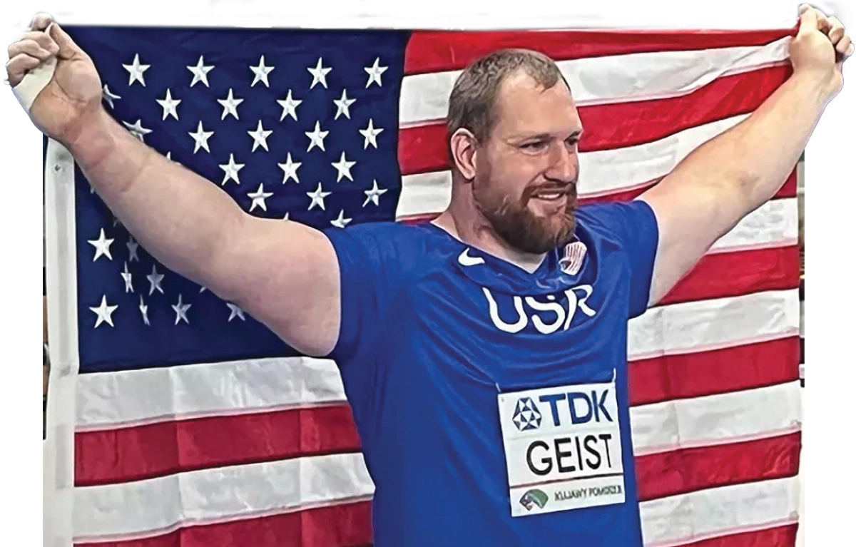 SRU assistant track and field coach Jordan Geist in a blue athletic t-shirt proudly smiling holding a United States of America flag behind him